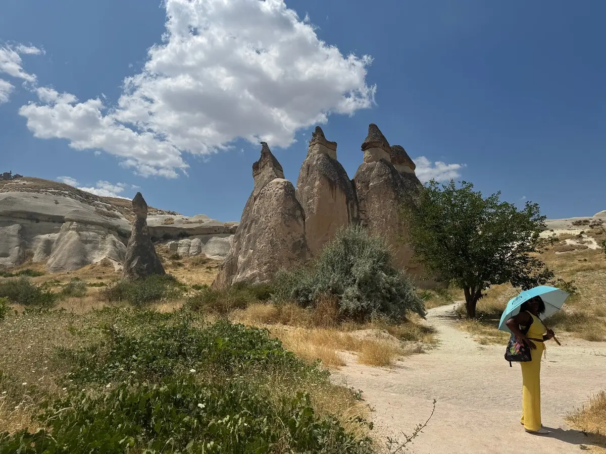 Un sendero de arena entre altas chimeneas de hadas cónicas bajo un cielo azul, con un visitante sosteniendo un paraguas